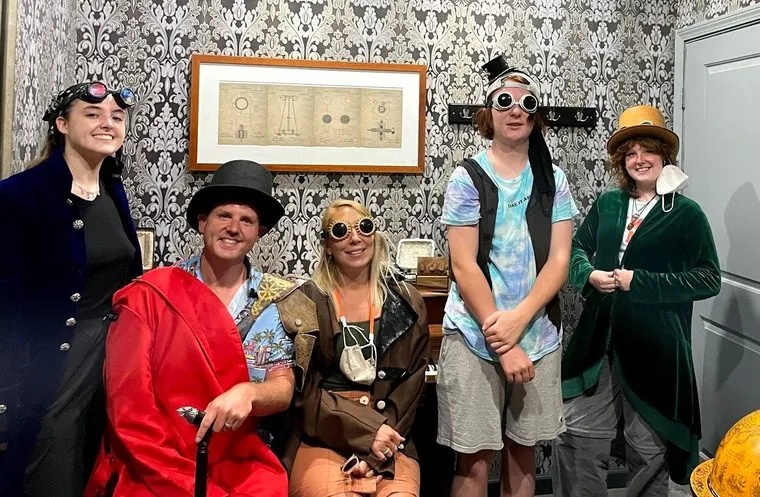 The family in steampunk costume — top hats, tinted goggles and long coats — against damask wallpaper, below a framed diagram of Victorian apparatus
